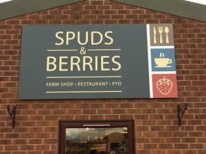 A black sign with gold lettering reading "Spuds & Berries Farm Shop - Restaurant - PYO" on the side of a red brick building.