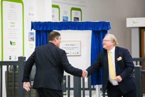 Two men in suits shaking hands in front of a plaque framed by curtains.
