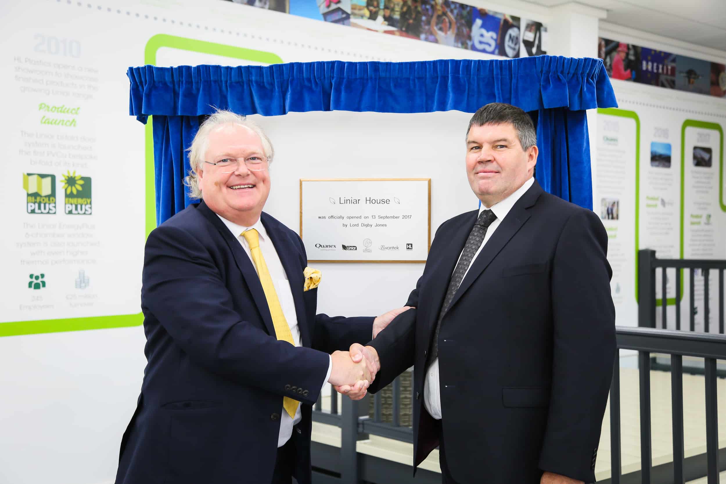 Two men in suits shaking hands in front of a plaque framed by curtains.