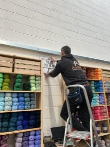 A wall of yarn, on which an Andrews-hoodie-clad man is installing a sign.