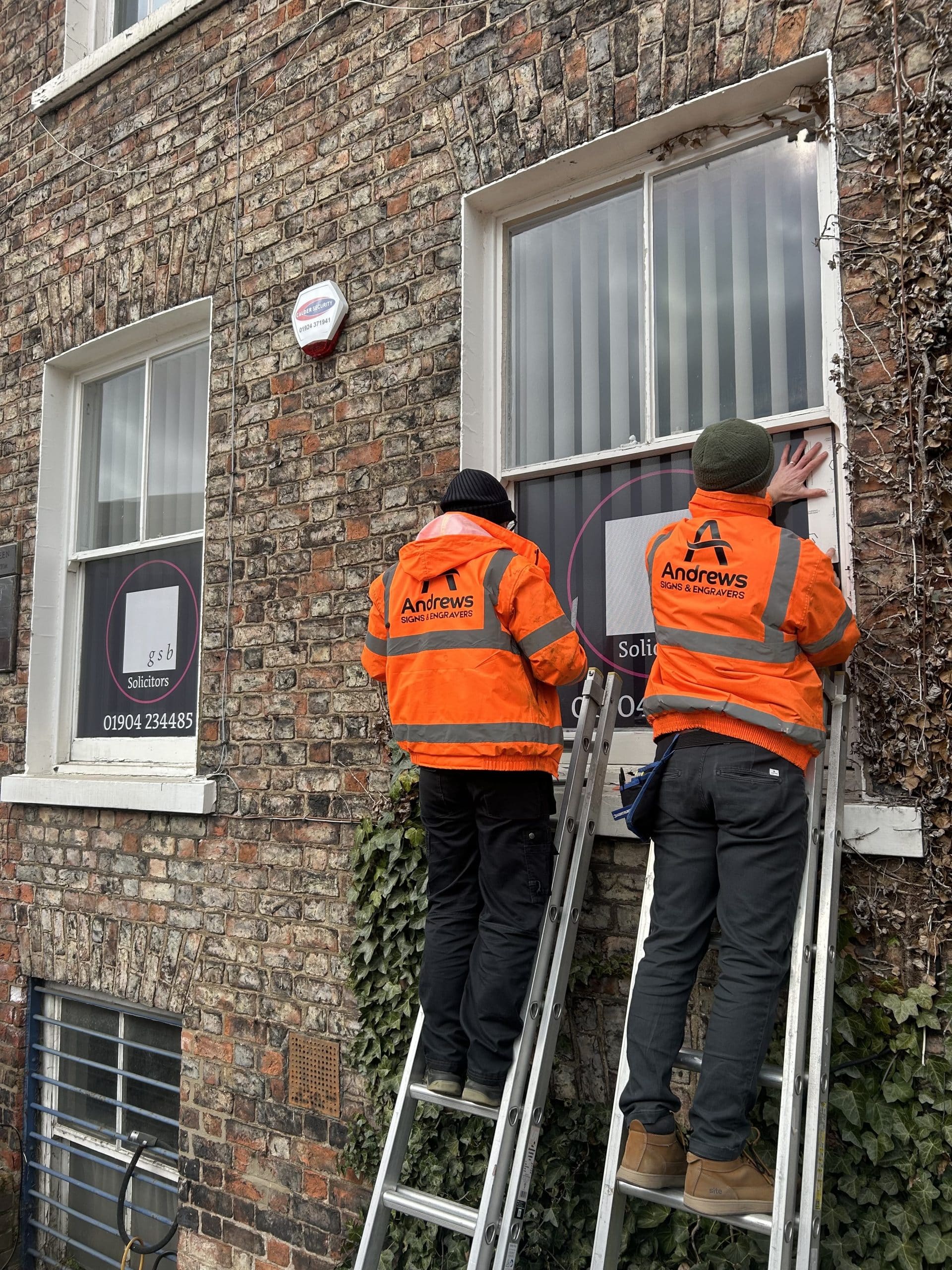 Two men in bright orange hi-vis coats standing on ladders applying a graphic to a window.