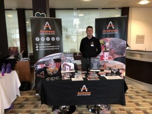 A man standing behind a table labeled Andrews, with lots of pamphlets on it.