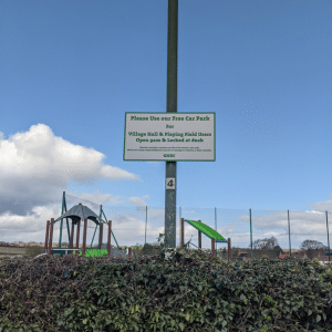 A white sign with green lettering saying "Please Use our Free Car Park", in front of a fenced-in playground.